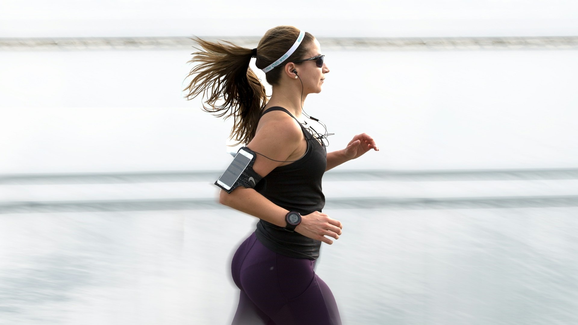 Vrouw jogt langs het water in paarse compressiekleding met sporthorloge en smartphonehouder